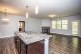 This single-floor residence is designed with an open-concept layout that connects the kitchen, dining, and living areas into one cohesive, well-lit space. The kitchen serves as a central hub, anchored by a large island with a double stainless steel sink and dark granite countertops. Rich, dark espresso cabinetry extends to the ceiling, providing generous storage, while black appliances — including a refrigerator, electric range, and microwave — offer a consistent, modern aesthetic. Pendant lighting above the island and a statement hanging fixture near the dining area add warmth and visual interest.

The living area features hardwood-style flooring that flows throughout the main living spaces, and a stone-surround fireplace serves as a focal point along one wall. Large windows allow natural light to fill the room, with views of greenery outside. A separate room with access to the exterior provides additional flexible living space, also finished with the same dark flooring.

The bedrooms are carpeted for comfort and include windows overlooking the outdoors. Two bathrooms serve the unit: one featuring a tub-shower combination, and the other a standalone glass-door shower with a dark vanity and white countertop — both finished with hardwood-style flooring consistent with the rest of the home.