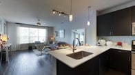 This single-floor space showcases a thoughtfully designed open-concept layout that seamlessly connects the kitchen and living areas. The kitchen is anchored by a spacious island with a crisp white countertop and an undermount stainless steel sink, complemented by dark cabinetry that creates a striking contrast. Overhead, a combination of industrial-style glass pendant lights and a track lighting system provides both focused task lighting and ambient illumination throughout the space.

The living area is bright and welcoming, benefiting from large windows dressed with sheer curtains that allow generous natural light to filter in. A ceiling fan adds both function and a subtle design element to the room. The wood-style flooring runs continuously throughout, unifying the open floor plan and adding warmth to the overall aesthetic.

The neutral color palette of soft grays and whites provides a versatile backdrop, while the contemporary furnishings and thoughtful decor accents contribute to a modern, polished ambiance. The seamless flow between the kitchen and living space makes this layout well-suited for both everyday living and entertaining.