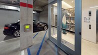 This single-level parking garage is designed with both functionality and modern convenience in mind. A standout feature is the dedicated row of wall-mounted electric vehicle charging stations, each equipped with illuminated blue connectors and individual cables, accommodating multiple EVs simultaneously. Overhead, an exposed ceiling reveals organized conduit runs, ventilation ducts, and utility infrastructure, lending the space an industrial yet well-maintained aesthetic.

Access to the garage is controlled via a keypad entry panel mounted at the entrance, ensuring secure ingress and egress. Adjacent to the parking area, a glass-enclosed surf and paddleboard storage room provides a distinctive amenity, housing an organized rack of boards alongside a decorative stand-up surfboard, catering to an active, outdoor lifestyle.

A glazed lobby enclosure connects the garage directly to the building's elevator system, branded with the "Sway" logo. This bright, cleanly finished lobby features polished flooring, warm wood wall panels, and a natural wood accent stool, creating a welcoming transition point between the parking level and the rest of the building. The combination of EV infrastructure, specialized storage, and a refined lobby entry reflects a thoughtfully designed, amenity-rich facility.