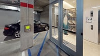 This single-level parking garage is designed with both functionality and modern convenience in mind. A standout feature is the dedicated row of wall-mounted electric vehicle charging stations, each equipped with illuminated blue connectors and individual cables, accommodating multiple EVs simultaneously. Overhead, an exposed ceiling reveals organized conduit runs, ventilation ducts, and utility infrastructure, lending the space an industrial yet well-maintained aesthetic.

Access to the garage is controlled via a keypad entry panel mounted at the entrance, ensuring secure ingress and egress. Adjacent to the parking area, a glass-enclosed surf and paddleboard storage room provides a distinctive amenity, housing an organized rack of boards alongside a decorative stand-up surfboard, catering to an active, outdoor lifestyle.

A glazed lobby enclosure connects the garage directly to the building's elevator system, branded with the "Sway" logo. This bright, cleanly finished lobby features polished flooring, warm wood wall panels, and a natural wood accent stool, creating a welcoming transition point between the parking level and the rest of the building. The combination of EV infrastructure, specialized storage, and a refined lobby entry reflects a thoughtfully designed, amenity-rich facility.