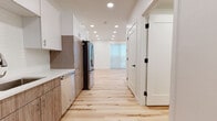 This single-floor unit showcases a clean, contemporary design throughout, unified by warm light hardwood flooring that runs seamlessly from room to room. The kitchen is a standout feature, outfitted with two-tone cabinetry — combining white upper cabinets with wood-grain lower cabinets — white quartz countertops, a subway tile backsplash, and a full suite of stainless steel appliances including a gas range, over-the-range microwave, dishwasher, and a French door refrigerator. Recessed lighting illuminates the kitchen and hallway, creating a bright and functional workspace.

The open living area is generously proportioned, bathed in natural light from a large sliding glass door that offers outdoor views and access to a balcony. Crisp white walls and recessed ceiling lights enhance the airy, minimalist ambiance of the space. The bedroom is a calm, neutral retreat with continued hardwood flooring and ample wall space.

The bathroom is thoughtfully finished with a wood-grain vanity with white countertop, a large frameless mirror, elegant pendant lighting, black hardware accents, and a full tub-shower combination. Together, these elements reflect a cohesive modern aesthetic that is both stylish and practical throughout the entire unit.