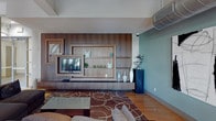 This thoughtfully designed single-floor space offers a sophisticated blend of contemporary style and functional versatility. The open-plan layout is anchored by warm hardwood flooring that flows seamlessly throughout, tying together distinct yet interconnected zones.

The main lounge area is a standout feature, centered around a custom built-in wood wall unit with integrated shelving and a media display, complemented by a plush sectional sofa and a patterned area rug. Bold teal accent walls provide a vibrant backdrop, while curated abstract artwork adds a gallery-like quality to the environment.

Adjacent to the lounge, a dining area accommodates multiple seating arrangements, including both round tables with upholstered chairs and bar-height seating. Large floor-to-ceiling grid-pane windows bathe this zone in natural light and provide views of lush outdoor greenery.

A well-appointed kitchenette serves the space with rich dark wood cabinetry, granite countertops, a glass tile backsplash, and a stainless steel under-counter refrigerator — ideal for hosting and entertaining. Floating shelves display decorative accessories that echo the teal-and-earth color palette found throughout.

Exposed industrial-style ductwork and track lighting on the ceiling add an urban, loft-inspired character, while structural columns define spaces without fully enclosing them, preserving the open and airy ambiance of the overall layout.