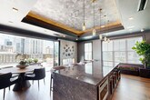 Located at 515 Royal Court in the heart of Charlotte, NC, this sophisticated amenity space delivers an elevated urban experience defined by refined design and premium finishes throughout. The interior is anchored by a dramatic tray ceiling adorned with a textured metallic surface and warm gold-leaf border, above which crystal pendant lights cast a polished glow over the space. A commanding marble island with a richly veined surface serves as the focal point of the main kitchen area, surrounded by upholstered bar seating and an adjacent dining nook. The second kitchen features floor-to-ceiling geometric metallic tile, dark cabinetry with brass hardware, marble countertops, open brass shelving, and stainless steel appliances — a striking blend of functionality and artistry. Large glass folding doors seamlessly connect the interior to an expansive outdoor terrace, where sweeping views of the Charlotte skyline take center stage. The rooftop terrace is equipped with an outdoor kitchen featuring a built-in grill and quartz countertop island, bar seating with an outdoor TV, string lighting, and curated planters — ideal for entertaining. Wide-plank wood-look flooring and large-format tile flow between the indoor and outdoor zones, creating a cohesive, luxurious environment throughout.