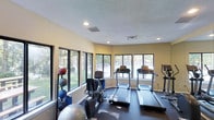 This single-floor fitness center offers a dedicated space for exercise and wellness. The room is lined with large, multi-pane windows along multiple walls, flooding the interior with natural light and providing pleasant views of the surrounding tree-lined outdoor environment. The textured ceiling features recessed lighting, supplementing the abundant daylight to keep the space well-lit at all times. The flooring is finished in a dark, durable material suited for gym use. The facility is equipped with a variety of cardio machines, including multiple treadmills and an elliptical trainer, positioned to take advantage of the outdoor views. A stationary bike and additional cardio equipment round out the machine offerings. Along the windowed walls, a dedicated stretching and functional training area is set up with an assortment of medicine balls and stability exercise balls of varying sizes, neatly organized on a multi-tier rack. The walls are finished in a soft, neutral tone that contributes to a clean and focused atmosphere. The overall layout is open and functional, allowing for ease of movement between equipment, making this fitness center a practical and inviting space for a full workout routine.
