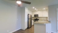 This single-floor residence offers a clean, modern aesthetic with thoughtful finishes throughout. The kitchen serves as a central highlight of the space, showcasing crisp white shaker-style cabinetry that extends to the ceiling for ample storage. Stainless steel appliances — including a full-size refrigerator, electric range, and built-in microwave — provide a sleek, contemporary look. Granite-style countertops add texture and elegance, while a kitchen island or peninsula offers additional workspace and casual dining potential. Recessed lighting illuminates the kitchen efficiently, complementing the warm hardwood-style flooring that runs throughout the space, adding both durability and visual warmth. The open layout connects the kitchen to the adjacent living area, creating a seamless flow ideal for everyday living and entertaining. A ceiling fan with integrated lighting enhances comfort and air circulation in the living area. Neutral wall tones in a soft gray-green palette create a calm, versatile backdrop that pairs well with a variety of décor styles. Overall, the space balances functional design with modern finishes, making it a comfortable and stylish single-floor home.