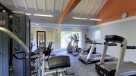 This fitness center offers a comprehensive workout environment designed to support a variety of exercise routines. The space is characterized by a striking vaulted ceiling with exposed white-painted beams accented by a bold orange fascia, lending the room a sense of height and openness. Flush-mounted fluorescent lighting ensures the interior remains bright throughout the day, complemented by abundant natural light streaming in through large sliding glass doors and side windows that frame lush greenery outside. The cardio section is well-appointed with multiple treadmills, elliptical trainers, and additional aerobic machines, all arranged to maximize space and usability. A dedicated strength training area features weight benches and cable resistance machines, providing options for a full-body workout. Large wall mirrors near the entrance enhance the sense of space while allowing users to monitor their form. The flooring consists of durable, patterned carpet tiles suited for a fitness environment. A glass-paned door provides a secondary access point, adding to the room's connectivity. Overall, the fitness center strikes a balance between functionality and comfort, offering an inviting atmosphere for both casual exercisers and dedicated fitness enthusiasts.