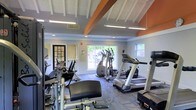 This fitness center offers a comprehensive workout environment designed to support a variety of exercise routines. The space is characterized by a striking vaulted ceiling with exposed white-painted beams accented by a bold orange fascia, lending the room a sense of height and openness. Flush-mounted fluorescent lighting ensures the interior remains bright throughout the day, complemented by abundant natural light streaming in through large sliding glass doors and side windows that frame lush greenery outside. The cardio section is well-appointed with multiple treadmills, elliptical trainers, and additional aerobic machines, all arranged to maximize space and usability. A dedicated strength training area features weight benches and cable resistance machines, providing options for a full-body workout. Large wall mirrors near the entrance enhance the sense of space while allowing users to monitor their form. The flooring consists of durable, patterned carpet tiles suited for a fitness environment. A glass-paned door provides a secondary access point, adding to the room's connectivity. Overall, the fitness center strikes a balance between functionality and comfort, offering an inviting atmosphere for both casual exercisers and dedicated fitness enthusiasts.