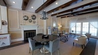 This single-floor space exudes refined elegance through a thoughtful blend of classic architectural details and carefully curated furnishings. The ceiling immediately draws the eye upward, where rich, dark-stained exposed wood beams stretch across the room, complemented by recessed lighting and a striking geometric chandelier that anchors the central dining area. Warm, neutral wall tones are enhanced by decorative wainscoting and paneling, lending the space a timeless, formal character.

A white-mantled fireplace serves as a focal point along one wall, adorned with decorative circular mirrors that add a contemporary touch to the traditional setting. The dining area features a dark round pedestal table surrounded by upholstered blue-gray chairs, offering an inviting space for gathering. Beyond, a comfortable lounge area is furnished with armchairs and geometric side tables, ideal for relaxed conversation.

Natural light filters in through large windows dressed in sheer white curtains, brightening the carpeted lounge zone and connecting the interior to the outdoors. A ceiling fan provides practical comfort overhead. Together, these elements combine to create a space that is both functional and visually distinguished, with an ambiance that balances warmth, elegance, and classic design sensibility.