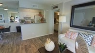 This single-story residence offers an open-concept floor plan that seamlessly connects the living area to the kitchen, creating a cohesive and functional living environment. The living space features warm wood-look flooring that extends throughout, complementing the neutral gray-toned walls and providing a clean, modern aesthetic.

The kitchen is positioned adjacent to the living area and is separated by a peninsula counter with a dark countertop, offering both a casual dining surface and a natural visual boundary between spaces. White cabinetry lines the kitchen walls, paired with white appliances, contributing to a bright and tidy appearance. A textured ceiling adds subtle architectural character to the space.

Visible beyond the main living area is a hallway leading to additional rooms, including what appears to be a bathroom with matching white cabinetry and light finishes, maintaining a consistent design language throughout. The layout is efficiently organized, allowing natural sightlines across the space. Overall, the residence presents a functional and well-coordinated interior with consistent finishes and a welcoming, casual ambiance suited for everyday living.