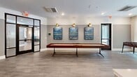 Located at 1300 Hendricks Ave in Jacksonville, Florida, this thoughtfully designed single-floor community space offers a sophisticated blend of social and leisure amenities. The open-concept layout is anchored by a striking elongated white quartz island, illuminated by a series of gold-accented glass pendant lights that add a warm, refined ambiance. Surrounding the island, charcoal barrel chairs with brushed gold legs provide stylish seating, while floor-to-ceiling windows bathe the kitchen and dining area in abundant natural light and frame lush, verdant outdoor views. A dedicated wet bar with dark cabinetry, marble-look backsplighting, and a utility sink sits adjacent to the island, completing a well-appointed entertaining kitchen. The living area features a curated lounge arrangement of curved, cream-upholstered chairs centered around a bold multi-panel video wall framed in warm wood, ideal for communal viewing. A full-length shuffleboard table anchors a game zone beneath a trio of framed artwork with brass wall sconces. A cozy banquette nook with dark cabinetry and bench seating offers an intimate gathering spot. Wide-plank wood-look tile flooring flows throughout, unifying the space with a clean, contemporary aesthetic.