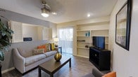 This single-floor residence offers a comfortable and well-appointed living space designed with both function and style in mind. The main living area serves as the heart of the home, featuring durable wood-look flooring that adds warmth and a contemporary feel. A ceiling fan with integrated lighting keeps the space comfortable year-round, while recessed lighting provides additional illumination. Built-in open shelving lines one wall, offering ample display and storage space alongside a dedicated area for entertainment setup. Natural light flows generously into the room through a large sliding glass door fitted with vertical blinds, which also provides convenient access to an outdoor area. The neutral color palette of warm beige and grey tones creates a calm, inviting ambiance that complements a variety of décor styles. A textured ceiling adds a classic touch to the overall interior. The layout is thoughtfully arranged to maximize the sense of openness, making the living area feel spacious and welcoming. Overall, this residence combines practical design elements with a bright, airy atmosphere to create a comfortable everyday living environment.