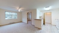 This single-floor unit offers a practical and well-organized layout centered around an open-concept living and kitchen area. The carpeted living room is generously sized, featuring a ceiling fan with light fixture and a large window that admits ample natural light while offering outdoor views of surrounding greenery. A baseboard heater runs beneath the window, providing efficient climate control. The kitchen is positioned adjacent to the living area, separated by a peninsula counter with white cabinetry above and below. White appliances, including a refrigerator and range, complement the tile flooring that extends through the kitchen and entry areas, creating a clean transition between spaces. The entry area includes a closet for additional storage. Two carpeted bedrooms each feature windows with blinds and baseboard heating, keeping the rooms bright and comfortable. The bathroom is notably spacious, finished with tile flooring, a long vanity with an extended countertop, ample cabinet storage with gold-toned hardware, a large mirror, a walk-in glass-enclosed shower, and wall-mounted shelving for added functionality. Throughout, the unit maintains a neutral, light color palette with warm wood-toned trim accents, creating a clean and cohesive aesthetic.