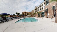Located at 211 River Landing Drive in Charleston, South Carolina, this outdoor amenity space at Simmons Park is designed to deliver a resort-like experience within a refined residential setting. The centerpiece of the space is a generously sized swimming pool, framed by a wide concrete deck lined with lounge chairs and shaded by navy umbrellas. A covered gazebo provides additional shade and gathering space poolside, while the surrounding multi-story brick building with green-trimmed balconies contributes to an upscale coastal aesthetic.

The covered outdoor lounge serves as a sophisticated social hub, featuring soaring coffered ceilings supported by exposed brick columns that lend architectural character throughout. Wicker seating arrangements with navy and teal accent pillows are arranged symmetrically on patterned stone tile flooring, creating an inviting atmosphere for relaxation and conversation. Wall-mounted flat-screen televisions and draped white curtains add to the cabana-like ambiance.

The lounge opens directly toward the pool area through teal-framed glass doors, creating a seamless indoor-outdoor flow. Additional features include a cornhole game area and built-in seating along the perimeter walkway. The overall design blends classic Southern brick craftsmanship with contemporary outdoor living amenities, making it an ideal gathering space for residents.