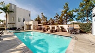 Located at 324 Parallel Gln in Escondido, California, this outdoor amenity space offers a resort-inspired pool and spa experience designed for relaxation and recreation. The centerpiece is a generously sized lap pool with crystal-clear blue water, framed by a broad sundeck outfitted with modern lounge chairs and umbrellas for shade. Adjacent to the pool sits a separate spa with bubbling jets, ideal for unwinding after a swim. Towering palm trees line the perimeter, lending a tropical ambiance that complements the sunny Southern California climate. The surrounding hardscape is clean and well-maintained, providing ample space for lounging and socializing. An outdoor fireplace and barbecue area are also present, making the space well-suited for gatherings. The backdrop features crisp, contemporary white-stucco multi-story buildings with clean architectural lines, reinforcing the modern aesthetic of the community. Lush shrubs and drought-tolerant plantings add greenery throughout, while the open layout ensures plenty of natural light and an airy, welcoming atmosphere. This thoughtfully designed outdoor space captures the essence of California outdoor living.