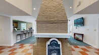 Located at 1700 Birch Trail Cir in Chesapeake, Virginia, this single-floor community clubhouse lounge offers a warm and inviting gathering space thoughtfully designed for both relaxation and socializing. The room's most striking architectural feature is its soaring vaulted ceiling, which lends a sense of grandeur and openness. A dramatic floor-to-ceiling stone fireplace anchors one side of the space, serving as a bold focal point and adding a cozy, residential character. Ceiling fans are positioned throughout to complement the volume of the high ceilings and maintain year-round comfort. The living area is furnished with comfortable seating, including a sofa, accent chairs, and a central coffee table, all arranged around the fireplace for an intimate atmosphere. An adjacent bar counter with barstool seating and a kitchenette behind it provides a convenient refreshment area. On the opposite end, a stunning floor-to-ceiling window wall curves around a café-style seating nook, flooding the interior with natural light and offering direct views of the outdoor pool and surrounding grounds. The flooring transitions between plush teal carpet in the lounge area and a bold checkered tile pattern near the entry. A wall-mounted flat-screen television adds a modern, functional touch to the space.