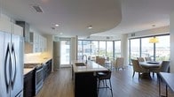 This single-floor space showcases a thoughtfully designed open-concept layout that seamlessly connects the kitchen and living areas. The kitchen is anchored by a substantial central island featuring a light quartz countertop, an undermount sink, and seating for multiple guests along its dark cabinetry base. Dark stainless steel appliances and white upper cabinetry line the kitchen wall, creating a striking contrast that adds visual interest.

A distinctive architectural ceiling detail — a curved, layered tray design — defines the transition between the kitchen and the living area, adding depth and character to the space. Pendant lighting hangs elegantly above the island, complementing the recessed lighting throughout.

The living area benefits from expansive floor-to-ceiling windows along multiple walls, flooding the interior with abundant natural light and offering broad views of the surrounding cityscape. Warm wood-tone flooring runs throughout the entire space, lending continuity and a welcoming ambiance. The overall aesthetic is contemporary and refined, balancing functional design with an open, airy atmosphere suited for both everyday living and entertaining.