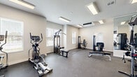 This fitness center in Rogers, Arkansas, offers a thoughtfully designed single-floor layout that accommodates a comprehensive range of exercise equipment. The space is organized into distinct functional zones, maximizing the available floor area for an efficient and comfortable workout experience.

The strength training area features a cable machine, multiple weight machines, a dumbbell rack with a full-length mirror wall, and a flat bench, providing ample options for resistance training. Adjacent to this zone, the cardio section houses two treadmills, an elliptical, a stationary spin bike, and a rowing machine, all positioned near windows that allow natural light to filter in.

The interior design is clean and modern, with neutral white walls accented by a gray shiplap feature wall. Durable rubber-speckled flooring spans the entire space, offering both safety and ease of maintenance. Recessed panel lighting and a wall-mounted split-system HVAC unit ensure a well-lit, climate-controlled environment year-round. A wall-mounted television in the cardio zone adds convenience for users during workouts.

A separate ancillary room with interior windows and a glass door is accessible from the main floor, providing an additional private area within the facility.