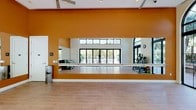 This single-floor fitness studio is thoughtfully designed to support a wide range of movement-based activities, from dance and barre classes to group fitness sessions. The space is immediately defined by its striking warm orange accent walls, which lend an energetic and motivating ambiance to the interior. A full-width wall mirror runs the length of one side of the room, paired with a mounted ballet barre—an essential feature for barre, dance, and stretching exercises alike. The mirror not only serves a functional purpose but also visually expands the sense of space within the studio. The flooring consists of smooth, wood-look planks that provide a clean, durable surface well-suited for athletic activities. Natural light flows generously into the studio through large floor-to-ceiling windows and glass doors, offering views of surrounding greenery and creating a bright, welcoming atmosphere. High ceilings with ceiling fans enhance airflow and contribute to the open, airy feel of the room. Additional fitness equipment and accessories, including colorful exercise balls, are visible along the perimeter, indicating the studio's versatility and readiness for varied workout programs.