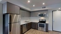 This single-floor space showcases a thoughtfully designed interior with a strong emphasis on modern aesthetics and functional living. The kitchen is a standout feature, outfitted with a full suite of stainless steel appliances including a refrigerator, electric range, over-the-range microwave, and dishwasher. High-gloss, two-toned cabinetry in a sleek gray finish lines the upper and lower walls, providing ample storage while contributing to the space's contemporary feel. The countertops are finished in a clean white surface that contrasts elegantly with the darker lower cabinets and wood-tone base cabinetry. A stainless steel undermount sink is positioned beneath recessed overhead lighting, which illuminates the space efficiently alongside a central HVAC vent for year-round comfort. The flooring throughout is a warm, light wood-look material that adds texture and warmth to the otherwise cool, modern palette. The open layout and recessed lighting create a bright, airy atmosphere. Overall, the space presents a clean, move-in-ready environment with a cohesive design language that balances practicality with contemporary style.