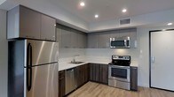 This single-floor space showcases a thoughtfully designed interior with a strong emphasis on modern aesthetics and functional living. The kitchen is a standout feature, outfitted with a full suite of stainless steel appliances including a refrigerator, electric range, over-the-range microwave, and dishwasher. High-gloss, two-toned cabinetry in a sleek gray finish lines the upper and lower walls, providing ample storage while contributing to the space's contemporary feel. The countertops are finished in a clean white surface that contrasts elegantly with the darker lower cabinets and wood-tone base cabinetry. A stainless steel undermount sink is positioned beneath recessed overhead lighting, which illuminates the space efficiently alongside a central HVAC vent for year-round comfort. The flooring throughout is a warm, light wood-look material that adds texture and warmth to the otherwise cool, modern palette. The open layout and recessed lighting create a bright, airy atmosphere. Overall, the space presents a clean, move-in-ready environment with a cohesive design language that balances practicality with contemporary style.