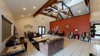 Located at 15095 Via Hondonada in San Diego, CA 92129, this single-story community clubhouse serves the Rancho Peñasquitos neighborhood with a welcoming and spacious interior designed for social gatherings and relaxation. The most striking architectural feature is the dramatic vaulted ceiling supported by exposed dark wood beam trusses, which soar overhead and lend a warm, lodge-inspired character to the main hall. Multiple ceiling fans are suspended from the trusses, ensuring comfortable airflow throughout the space. The main gathering area is generously proportioned, finished with large-format white ceramic tile flooring that reflects natural light and enhances the sense of openness. Bold terracotta-red accent walls provide visual contrast against the crisp white walls and ceiling, creating a vibrant yet inviting ambiance. The seating area is furnished with plush sofas and accent chairs arranged around coffee tables, making it ideal for casual conversation or community events. Additional seating is available at dining tables and chairs positioned throughout the room. French doors on two sides of the hall allow natural light to flood the interior and provide access to the outdoor areas beyond. Recessed lighting and wall-mounted lamps supplement natural light for evening use. A water cooler station and a step-up platform area offer added functionality for community programming and events.