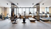 Located at 1114 Rue Saint-Antoine Ouest in Montréal, this professionally appointed fitness center offers a generously sized, single-floor exercise facility designed with both functionality and aesthetics in mind. The space features a clean, contemporary design characterized by large-format tile and rubber flooring, white ceilings with recessed and surface-mounted spotlights, and distinctive black-framed mirrored columns that visually expand the room and enhance the sense of depth throughout.

Floor-to-ceiling windows line the perimeter, flooding the space with natural light and offering sweeping views of the Montréal skyline — a motivating backdrop for any workout. The cardio zone is well-equipped with a row of treadmills, elliptical trainers, and stationary bikes, all from the premium Technogym brand. A dedicated strength training area houses an extensive selection of cable machines, multi-function weight stations, and benches, while a full free-weights section features a complete rack of dumbbells paired with adjustable benches and mirrored walls.

A separate, glass-enclosed room — visible from the main floor — provides a quieter space for stretching or ancillary training. The layout flows intuitively from cardio to strength zones, making the facility suitable for a wide range of fitness routines. The overall ambiance is polished and professional, befitting a high-end urban fitness facility.