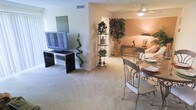 This single-floor residence offers a seamlessly connected living and dining layout, creating an open and functional environment. The living area is anchored by plush, light-toned carpeting that extends throughout, contributing to a warm and comfortable ambiance. A flat-screen television rests on a sleek, dark media console positioned against a crisp white wall, while a decorative wrought iron etagere displays an assortment of houseplants, adding natural character to the space.

The dining area features an elegant round glass-top table accompanied by ornate metal chairs, providing a refined spot for meals. Overhead, a ceiling fan with integrated lighting serves both the dining and adjacent sitting areas, ensuring comfort and illumination throughout.

The living room transitions smoothly into a cozy sitting nook warmed by soft ambient lighting. A large decorative tree plant anchors this corner, complementing a console table adorned with tasteful accessories. Warm peach-toned walls in this area create a welcoming contrast to the lighter tones of the main living space. Overall, the layout balances practicality with a welcoming, homey aesthetic suited to everyday living and entertaining.