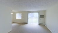 This single-floor space features a generously sized living area finished in a clean, neutral palette. Cream-colored walls and light wall-to-wall carpeting create a bright, airy atmosphere throughout the room. A large sliding glass door serves as the focal point of the main living area, fitted with vertical blinds and offering a direct view of a green outdoor area, allowing natural light to flood the interior. A secondary window on the adjacent wall further enhances the sense of brightness and openness. Baseboard heating units line the lower portion of the walls, providing an efficient and unobtrusive heating solution, while a wall-mounted air conditioning unit ensures year-round climate control. The textured ceiling adds a subtle visual element that complements the overall simplicity of the space. The layout is open and versatile, providing ample room for a variety of furniture arrangements. The overall ambiance is clean, functional, and welcoming, making it well-suited for comfortable everyday living.