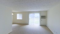 This single-floor space features a generously sized living area finished in a clean, neutral palette. Cream-colored walls and light wall-to-wall carpeting create a bright, airy atmosphere throughout the room. A large sliding glass door serves as the focal point of the main living area, fitted with vertical blinds and offering a direct view of a green outdoor area, allowing natural light to flood the interior. A secondary window on the adjacent wall further enhances the sense of brightness and openness. Baseboard heating units line the lower portion of the walls, providing an efficient and unobtrusive heating solution, while a wall-mounted air conditioning unit ensures year-round climate control. The textured ceiling adds a subtle visual element that complements the overall simplicity of the space. The layout is open and versatile, providing ample room for a variety of furniture arrangements. The overall ambiance is clean, functional, and welcoming, making it well-suited for comfortable everyday living.