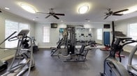 This fitness center offers a comprehensive workout environment designed to support a wide range of exercise routines. The space is laid out on a single floor with rubber flooring throughout, providing a durable and slip-resistant surface suited for high-activity use. Crown molding and soft, neutral-toned walls lend a polished, well-maintained aesthetic to the facility.

The cardio area is equipped with multiple treadmills, elliptical trainers, and a stair climber, positioned to take advantage of natural light from several windows fitted with blinds. A wall-mounted flat-screen TV is available for entertainment during workouts. Ceiling fans and recessed lighting keep the environment comfortable and well-illuminated.

The strength training section features a multi-station cable machine with various attachment options, a dedicated adjustable weight bench, and a full dumbbell rack spanning a range of weights. A large wall mirror spans a significant portion of the space, enhancing the sense of openness and allowing for proper form checks during exercise.

Additional equipment includes an upright exercise bike, stability and medicine balls displayed on a mounted rack, and a padded exercise mat for stretching or floor work. The facility is accessed through a glass-paneled door that provides a view of the surrounding outdoor greenery, contributing to an open and inviting atmosphere.
