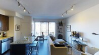 This single-floor residence showcases an open-concept layout that seamlessly connects the kitchen, dining, and living areas into one cohesive space. The kitchen features a dark-toned island with granite countertops and bar seating, providing both a functional workspace and a casual dining option. Adjacent to the kitchen, a traditional wooden dining table with classic chairs creates a defined yet integrated dining area.

The living space is furnished with a neutral-toned sofa and accent chairs, centered around a media console and complemented by a tall metal shelving unit displaying curated décor. Track lighting runs along the ceiling, providing adjustable illumination throughout the open space. A large sliding glass door floods the interior with natural light and opens to a balcony that overlooks the surrounding urban landscape, offering views of neighboring buildings. The balcony also accommodates outdoor seating, extending the living area outdoors. The overall ambiance is modern and well-appointed, balancing practical design with stylish, eclectic furnishings.