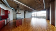 This single-floor urban loft showcases a bold industrial-modern aesthetic defined by exposed concrete ceilings with visible ductwork, warm hardwood flooring, and dramatic floor-to-ceiling windows that flood the open living area with natural light and frame expansive city and treetop views. The generous open-plan living and dining area flows seamlessly from a central kitchen island topped with a dark granite surface and fitted with a stainless steel ventilation hood. The full kitchen features rich, deep-red cabinetry with glass-front upper doors, dark granite countertops, a dark tile backsplash, and a complete suite of stainless steel appliances including a French door refrigerator, double wall ovens, built-in microwave, dishwasher, and cooktop. Track and pendant lighting throughout complement the industrial character of the space. The bedrooms are carpeted for comfort, with one benefiting from its own floor-to-ceiling windows and an exposed concrete column, bringing in abundant natural light. The bathrooms are finished with large-format neutral tile, including a soaking tub with tiled surround accented by a decorative mosaic border, and brushed nickel fixtures. A private balcony accessible through sliding glass doors offers elevated views of the surrounding cityscape.