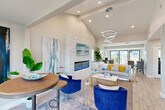 Located at 8135 N 34th Ave in Phoenix, Arizona, this single-floor office space delivers a polished and welcoming environment designed to impress. The layout is anchored by a spacious lounge area featuring soaring vaulted ceilings with exposed beams and a dramatic stacked-stone accent wall housing a sleek linear electric fireplace topped with a flat-screen display. A sculptural multi-ring chandelier adds a contemporary focal point overhead, while recessed lighting throughout ensures a bright, inviting atmosphere. The lounge is furnished with a plush neutral sofa accented by vibrant yellow cushions, bold cobalt blue accent chairs, and a marble-top coffee table resting on a patterned area rug over light wood-look flooring. Large-scale coastal artwork and lush indoor plants reinforce the fresh, coastal-inspired aesthetic. Transitioning to the office area, floor-to-ceiling black-framed windows and sliding glass doors flood the workspace with natural light and offer open views of the landscaped exterior grounds. White glass-top desks, upholstered chairs, and organized workstations create a clean, functional environment for staff. The overall design seamlessly blends comfort with professionalism, making this Phoenix office space both highly functional and visually distinctive.