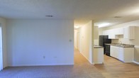 This single-story residence offers a straightforward and functional layout designed for comfortable everyday living. The living area is finished with wall-to-wall carpeting and neutral-toned walls, creating a clean and versatile canvas for furnishing and personalization. A textured ceiling adds a subtle architectural detail throughout the space, while ceiling-mounted light fixtures provide consistent overhead illumination.

Transitioning from the living area, a central hallway extends toward the private areas of the home, with doors visible along its length suggesting access to bedrooms or additional rooms. The hallway maintains the same neutral color palette, contributing to a cohesive feel throughout.

Adjacent to the living space, the kitchen is partially open to the main area via a countertop bar or pass-through peninsula, promoting a connected feel between the two spaces. The kitchen is equipped with a full-size black refrigerator, and the flooring transitions from carpet to hard surface, likely tile or vinyl, appropriate for a kitchen environment. White cabinetry lines the upper walls, and the dark countertops provide a contrasting accent. The layout appears practical and efficiently organized for everyday meal preparation.