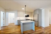 Located at 873 Communipaw Avenue in Jersey City, NJ, this well-appointed 2-bedroom, 2-bathroom residence offers a thoughtfully designed single-floor layout with modern finishes throughout. Warm hardwood-style flooring flows seamlessly across the entire space, complementing the clean white walls and contemporary aesthetic.

The open-concept kitchen and living area serves as the heart of the home. The kitchen is outfitted with sleek gray flat-front cabinetry extending to the ceiling, a white tile backsplash, stainless steel appliances including a refrigerator, range, dishwasher, and microwave, plus a central island with a white countertop and a stylish pendant light overhead. The spacious living area is bathed in natural light and opens directly to a private balcony through large floor-to-ceiling glass doors with black framing.

The generously sized primary bedroom features a wall-mounted split HVAC unit and ample closet space. Three closets in total provide excellent storage throughout the residence. A dedicated den offers flexible use as a home office or additional living space. In-unit laundry adds everyday convenience.

Both bathrooms are finished with modern tile work. One features a glass-enclosed walk-in shower with large-format tile, while the other showcases a vessel sink vanity with mirrored upper cabinetry and vertical subway tile accents.
