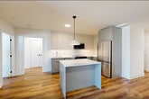 Located at 873 Communipaw Avenue in Jersey City, NJ, this well-appointed 2-bedroom, 2-bathroom residence offers a thoughtfully designed single-floor layout with modern finishes throughout. Warm hardwood-style flooring flows seamlessly across the entire space, complementing the clean white walls and contemporary aesthetic.

The open-concept kitchen and living area serves as the heart of the home. The kitchen is outfitted with sleek gray flat-front cabinetry extending to the ceiling, a white tile backsplash, stainless steel appliances including a refrigerator, range, dishwasher, and microwave, plus a central island with a white countertop and a stylish pendant light overhead. The spacious living area is bathed in natural light and opens directly to a private balcony through large floor-to-ceiling glass doors with black framing.

The generously sized primary bedroom features a wall-mounted split HVAC unit and ample closet space. Three closets in total provide excellent storage throughout the residence. A dedicated den offers flexible use as a home office or additional living space. In-unit laundry adds everyday convenience.

Both bathrooms are finished with modern tile work. One features a glass-enclosed walk-in shower with large-format tile, while the other showcases a vessel sink vanity with mirrored upper cabinetry and vertical subway tile accents.