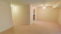 This single-floor residential space offers a clean, functional layout designed for comfortable everyday living. The main living area is generously proportioned, featuring wall-to-wall carpeting in a warm, neutral tone that adds softness underfoot and contributes to the overall understated aesthetic. Smooth, cream-colored walls provide a bright and airy backdrop throughout, while a ceiling fan with integrated lighting ensures year-round comfort and ambient illumination. A partial dividing wall creates a subtle sense of separation within the open floor plan, adding architectural interest without compromising the spacious feel of the area. A dedicated thermostat panel is conveniently positioned on the wall, indicating climate control throughout the unit. Adjacent to the living space, the kitchen is accessible through an open doorway and is outfitted with warm wood-toned cabinetry that provides ample storage. The kitchen also features dark countertops and essential appliances, making it a practical and efficient space for meal preparation. The overall ambiance of the space is calm and neutral, offering a versatile canvas that can be easily personalized to suit a variety of styles and preferences.