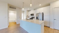 This single-story, one-bedroom, one-bathroom unit is designed with a clean, modern aesthetic highlighted by a neutral color palette and an abundance of natural light. The open-concept layout connects the living area to the kitchen seamlessly, creating a functional and cohesive living space. Light-toned wood-style flooring extends throughout the main areas, lending warmth and continuity to the design.

The kitchen serves as a central feature, equipped with stainless steel appliances including a French door refrigerator, electric range, built-in microwave, and dishwasher. White shaker-style cabinetry reaches toward the ceiling, maximizing storage, while a subway tile backsplash adds a refined detail. A large island with a quartz countertop and undermount sink provides additional workspace and casual seating potential. Pendant lighting above the island complements the recessed lighting throughout.

The living area offers generous wall space and large windows dressed with blinds, allowing for controlled natural light. The bedroom is carpeted for comfort, featuring multiple windows, a ceiling fan, and ample natural brightness. A dedicated laundry room houses a full-size washer and dryer with overhead shelving and a window. The bathroom is accessible and well-appointed, completing this thoughtfully designed unit.