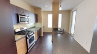 This single-floor space is centered around a well-appointed kitchen that seamlessly connects to an open living area. The kitchen is outfitted with stainless steel appliances, including a gas range, oven, and over-the-range microwave, all complemented by rich dark wood cabinetry that extends to the upper walls for ample storage. Granite-style countertops in muted gray tones run along both sides of the kitchen, with one side featuring a deep undermount sink paired with a modern faucet. A pendant light fixture adds a refined touch above the counter space. The flooring throughout is a dark hardwood, creating a warm and grounded aesthetic that contrasts cleanly with the soft, neutral yellow-toned walls. Natural light flows generously into the space through multiple windows fitted with horizontal blinds, illuminating both the kitchen and the adjacent living area. The open layout allows for a natural flow between cooking and living spaces, making the overall environment feel functional, modern, and bright.
