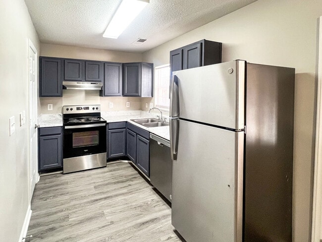 Open kitchen with stainless steel refrigerator, electric range, dishwasher, gray cabinets, marble style counters, and wood look flooring - Pendleton Townhomes