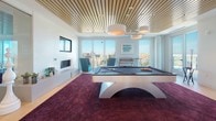 This contemporary amenity lounge is thoughtfully designed to offer both entertainment and relaxation in a sophisticated setting. The centerpiece of the space is a sleek, modern billiard table positioned atop a rich, deep-purple textured area rug, creating a bold visual anchor against the warm light hardwood flooring that extends throughout. Overhead, a distinctive slatted wood ceiling adds warmth and architectural interest, complemented by stylish drum pendant lighting.

Floor-to-ceiling windows line the perimeter, flooding the interior with abundant natural light and framing sweeping panoramic city views. A wall-mounted flat-screen television sits above a low-profile linear fireplace, flanked by vibrant teal accent chairs and additional seating that create an inviting lounge area. Decorative artwork and carefully curated furnishings contribute to the space's polished, upscale aesthetic.

Frameless glass sliding doors partition a separate corridor zone, maintaining an open, airy feel while defining distinct areas within the space. A sliding glass door leads directly to an outdoor terrace, where additional seating is visible, seamlessly connecting the interior to the exterior. The overall ambiance balances modern design with comfortable livability, making it an ideal space for socializing and recreation.