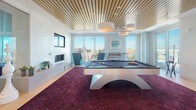 This contemporary amenity lounge is thoughtfully designed to offer both entertainment and relaxation in a sophisticated setting. The centerpiece of the space is a sleek, modern billiard table positioned atop a rich, deep-purple textured area rug, creating a bold visual anchor against the warm light hardwood flooring that extends throughout. Overhead, a distinctive slatted wood ceiling adds warmth and architectural interest, complemented by stylish drum pendant lighting.

Floor-to-ceiling windows line the perimeter, flooding the interior with abundant natural light and framing sweeping panoramic city views. A wall-mounted flat-screen television sits above a low-profile linear fireplace, flanked by vibrant teal accent chairs and additional seating that create an inviting lounge area. Decorative artwork and carefully curated furnishings contribute to the space's polished, upscale aesthetic.

Frameless glass sliding doors partition a separate corridor zone, maintaining an open, airy feel while defining distinct areas within the space. A sliding glass door leads directly to an outdoor terrace, where additional seating is visible, seamlessly connecting the interior to the exterior. The overall ambiance balances modern design with comfortable livability, making it an ideal space for socializing and recreation.