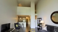 This two-story space makes a striking first impression with its soaring double-height ceilings that flood the main living area with a sense of openness and volume. The open-concept design connects the living room and kitchen in a fluid, cohesive layout, ideal for both everyday living and entertaining. A bold structural column anchors the space visually while delineating zones without interrupting the flow.

The kitchen features dark countertops atop a breakfast bar with bar stool seating, complemented by pendant lighting that adds a warm, modern touch. White upper cabinetry and stainless steel appliances lend a clean, contemporary aesthetic. Dark hardwood flooring runs throughout the main level, grounding the space and providing a rich contrast to the light-toned walls.

The upper level is visible from below, with an open mezzanine-style overlook that reinforces the vertical drama of the space. A shelving unit and accent lamp in the living area add functional character. Overall, the space conveys a modern, urban aesthetic with an emphasis on architectural detail, scale, and thoughtful design.