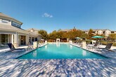 Located at 215 Republic Ave in Lafayette, Louisiana, this outdoor amenity space centers around a generously sized, resort-style swimming pool with crystal-clear blue water. The pool is designed with gradual entry points and classic stainless steel handrails, making it accessible and inviting. Surrounding the pool is a wide, well-maintained concrete sundeck furnished with an array of reclining lounge chairs and teal-accented umbrellas that provide comfortable shaded seating throughout the day. Stone and masonry accent columns frame the pool area, adding a refined architectural detail to the outdoor environment. An outdoor fireplace feature toward the rear of the deck offers a gathering focal point beyond the pool. The perimeter of the space is enclosed by a black wrought-iron fence, ensuring privacy and security, while mature trees and carefully maintained landscaping create a natural buffer and add to the serene atmosphere. A clubhouse-style building with a pitched roof, large windows, and covered porch elements sits adjacent to the pool, providing direct access to interior amenities. Under abundant natural sunlight and open skies, this outdoor space offers a well-appointed, community-oriented retreat ideally suited for relaxation and recreation in the heart of Lafayette.
