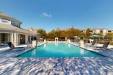 Located at 215 Republic Ave in Lafayette, Louisiana, this outdoor amenity space centers around a generously sized, resort-style swimming pool with crystal-clear blue water. The pool is designed with gradual entry points and classic stainless steel handrails, making it accessible and inviting. Surrounding the pool is a wide, well-maintained concrete sundeck furnished with an array of reclining lounge chairs and teal-accented umbrellas that provide comfortable shaded seating throughout the day. Stone and masonry accent columns frame the pool area, adding a refined architectural detail to the outdoor environment. An outdoor fireplace feature toward the rear of the deck offers a gathering focal point beyond the pool. The perimeter of the space is enclosed by a black wrought-iron fence, ensuring privacy and security, while mature trees and carefully maintained landscaping create a natural buffer and add to the serene atmosphere. A clubhouse-style building with a pitched roof, large windows, and covered porch elements sits adjacent to the pool, providing direct access to interior amenities. Under abundant natural sunlight and open skies, this outdoor space offers a well-appointed, community-oriented retreat ideally suited for relaxation and recreation in the heart of Lafayette.