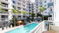 This outdoor amenity space is centered around a generously sized swimming pool with clear turquoise water, framed by a well-maintained courtyard setting. The pool area is surrounded by a wide deck furnished with lounge chairs and chaise seating, providing ample space for relaxation. A distinctive canopied gazebo with warm yellow fabric anchors one corner of the courtyard, offering a shaded retreat.

Tall, mature palm trees are strategically placed throughout the space, lending a tropical character and a sense of established landscaping. Additional umbrellas and lounge seating extend along the pool's perimeter, enhancing the resort-like atmosphere.

The courtyard is enclosed on multiple sides by a multi-story building with a crisp white exterior. Each floor features private balconies with white railings, many of which look directly down onto the pool area, creating an inward-facing, community-oriented design. Large windows accompany each balcony, suggesting bright, well-lit interiors.

The overall ambiance is bright and airy, evoking a warm-weather, leisure-focused environment with a clean, contemporary aesthetic suited to an active, outdoor-oriented lifestyle.