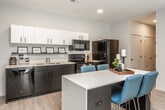 This single-floor unit showcases a contemporary design with a clean, neutral color palette and light wood-style flooring carried throughout. The open-concept kitchen is a standout feature, equipped with a full suite of black appliances — including a French door refrigerator, electric range, over-the-range microwave, and dishwasher. Two-tone cabinetry pairs white upper cabinets with dark lower cabinets, complemented by light countertops. A kitchen island with seating for multiple provides additional workspace and a casual dining option.

The living area is well-proportioned and bathed in natural light from sliding glass doors that open to a private balcony. The space offers a comfortable layout suitable for both relaxing and entertaining. The bedroom is thoughtfully sized, featuring a window with exterior views and room for a bed and dedicated study area.

Two bathrooms serve the unit, each consistently finished with dark vanity cabinetry, white countertops, framed mirrors, and a shower-tub combination. Recessed lighting throughout and a smart thermostat contribute to the unit's modern, functional character.