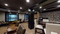 This single-floor space is designed as a multifunctional amenity lounge that balances social, productive, and casual uses within a cohesive, contemporary aesthetic. The interior is defined by a bold contrast of dark-painted ceilings with recessed tray detailing and dark structural columns set against warm beige walls and travertine-style tile flooring that flows throughout.

The lounge area features textured leather seating arranged around low tables, with a large framed Scrabble board mounted on the wall serving as a distinctive decorative focal point. Large dark-framed windows allow natural light to filter into the space while offering exterior views, complementing the ambient recessed lighting overhead.

A kitchenette runs along one wall, finished with dark cabinetry, a mosaic tile backsplash, countertop appliances, and a refrigerator, making it well-suited for light refreshments. A high-top dining table with white upholstered bar stools provides additional seating nearby.

A dedicated workspace alcove features a curved granite-topped desk accommodating two workstations, paired with ergonomic task chairs and a large corkboard panel framed in dark trim for notes and materials. Decorative sculptural elements and greenery are placed throughout, adding warmth and character to the overall environment.
