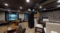 This single-floor space is designed as a multifunctional amenity lounge that balances social, productive, and casual uses within a cohesive, contemporary aesthetic. The interior is defined by a bold contrast of dark-painted ceilings with recessed tray detailing and dark structural columns set against warm beige walls and travertine-style tile flooring that flows throughout.

The lounge area features textured leather seating arranged around low tables, with a large framed Scrabble board mounted on the wall serving as a distinctive decorative focal point. Large dark-framed windows allow natural light to filter into the space while offering exterior views, complementing the ambient recessed lighting overhead.

A kitchenette runs along one wall, finished with dark cabinetry, a mosaic tile backsplash, countertop appliances, and a refrigerator, making it well-suited for light refreshments. A high-top dining table with white upholstered bar stools provides additional seating nearby.

A dedicated workspace alcove features a curved granite-topped desk accommodating two workstations, paired with ergonomic task chairs and a large corkboard panel framed in dark trim for notes and materials. Decorative sculptural elements and greenery are placed throughout, adding warmth and character to the overall environment.