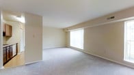 This single-floor space presents a clean, understated interior designed for comfortable everyday living. The main living area is generously proportioned, offering an open layout that provides flexibility for a variety of furniture arrangements. Soft, neutral beige walls create a warm and inviting atmosphere throughout, complementing the plush wall-to-wall carpeting underfoot, which adds a layer of comfort to the space. A large window serves as a focal point along one wall, allowing an abundance of natural light to filter into the room and contributing to an airy, bright ambiance. White baseboards provide a crisp contrast against the neutral walls, adding a subtle architectural detail that defines the room's perimeter. The ceiling is smooth and unadorned, maintaining the space's clean aesthetic. A wall-mounted light switch panel is visible, indicating practical, accessible electrical placement. The overall design is simple and functional, offering a blank canvas that can be easily personalized to suit a range of interior styles and preferences.
